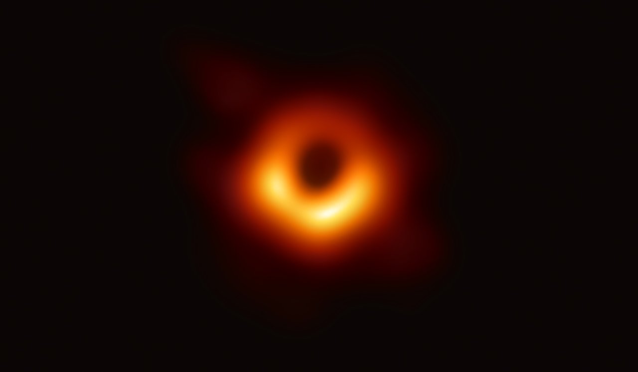 Oficjalne zdjęcie czarnej dziury. Czarna, okrągła plama otoczona rozmytą, pomarańczową otoczką o różnych odcieniach, na czarnym tle